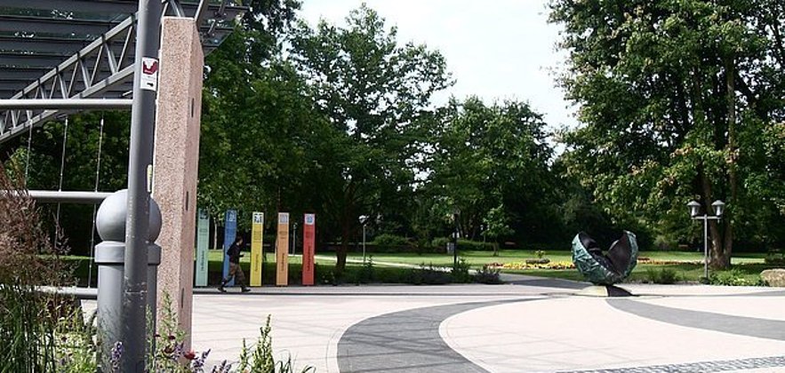Treffpunkt ist am Eingang zum Kurpark Bad Bergzabern. Foto: Tourismusverein Bad Bergzabern e.V. Blick auf einen modern gestalteten Platz mit geschwungenen Pflasterflächen, bunten Informationsstelen und einer Skulptur. Bäume und Grünflächen erstrecken sich dahinter.