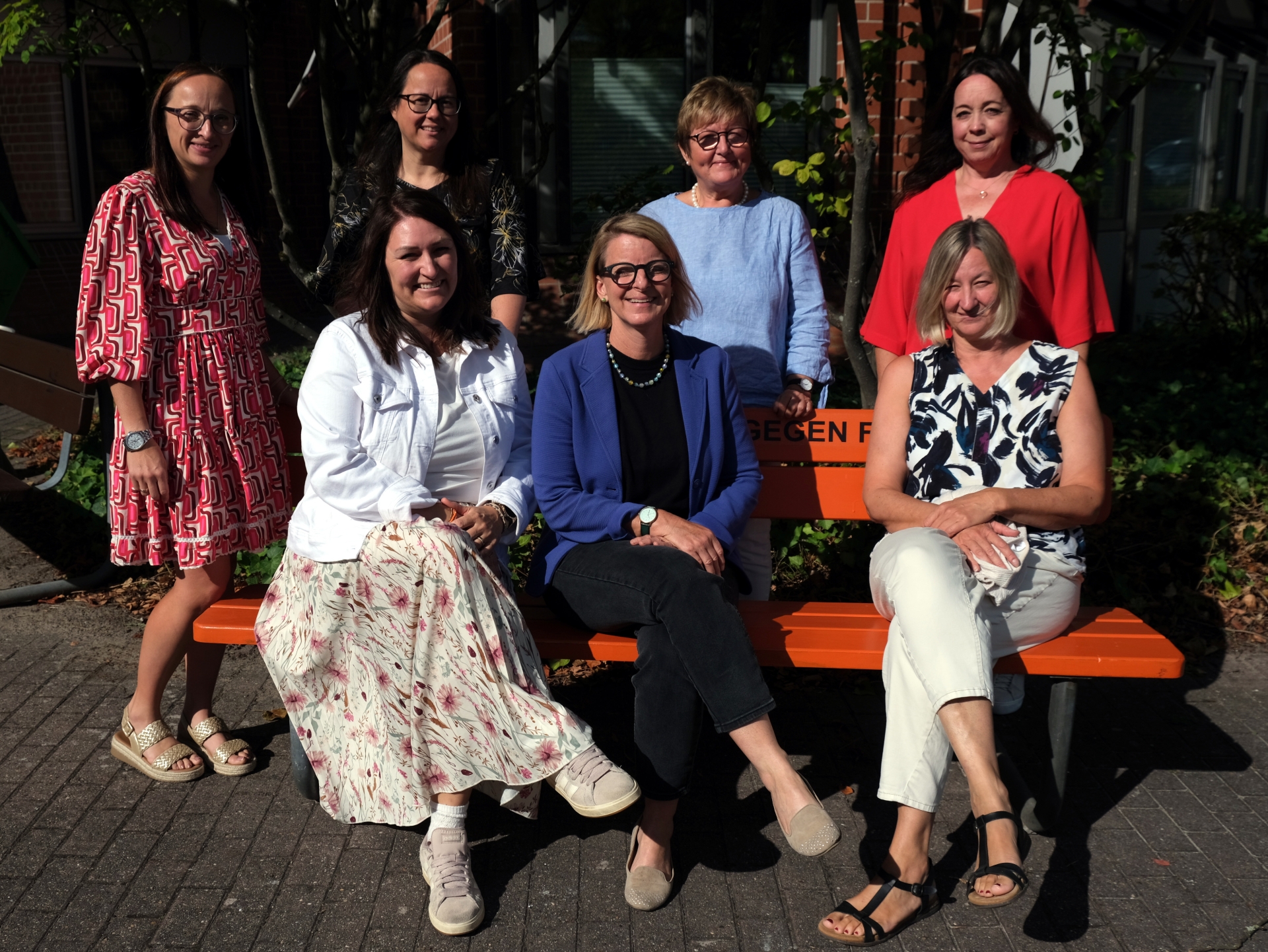 Sieben Frauen stehen und sitzen für ein Gruppenfoto bei Sonnenschein vor einem Gebäude. Drei von ihnen sitzen auf einer orangefarbenen Bank, vier stehen dahinter