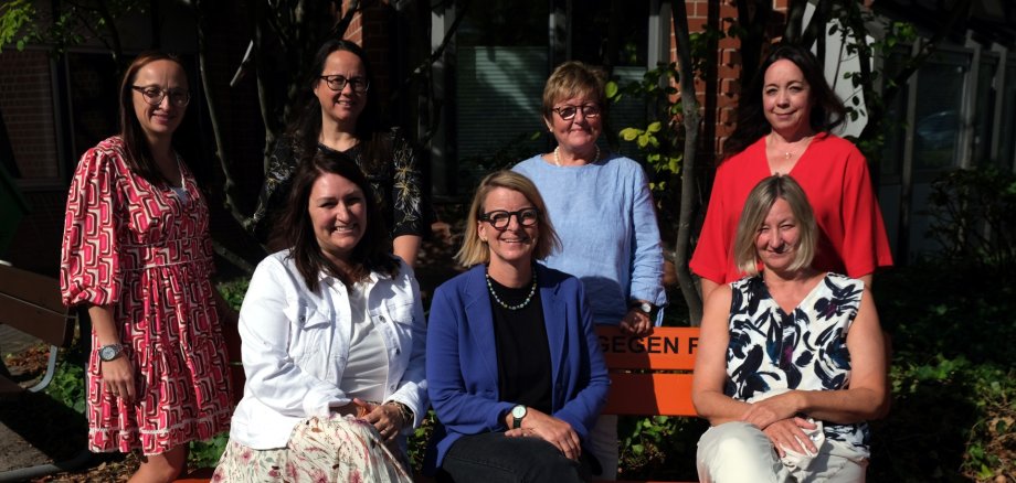 Sieben Frauen stehen und sitzen für ein Gruppenfoto bei Sonnenschein vor einem Gebäude. Drei von ihnen sitzen auf einer orangefarbenen Bank, vier stehen dahinter