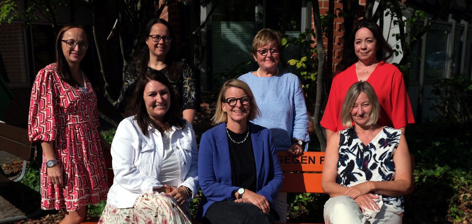 Sieben Frauen stehen und sitzen für ein Gruppenfoto bei Sonnenschein vor einem Gebäude. Drei von ihnen sitzen auf einer orangefarbenen Bank, vier stehen dahinter