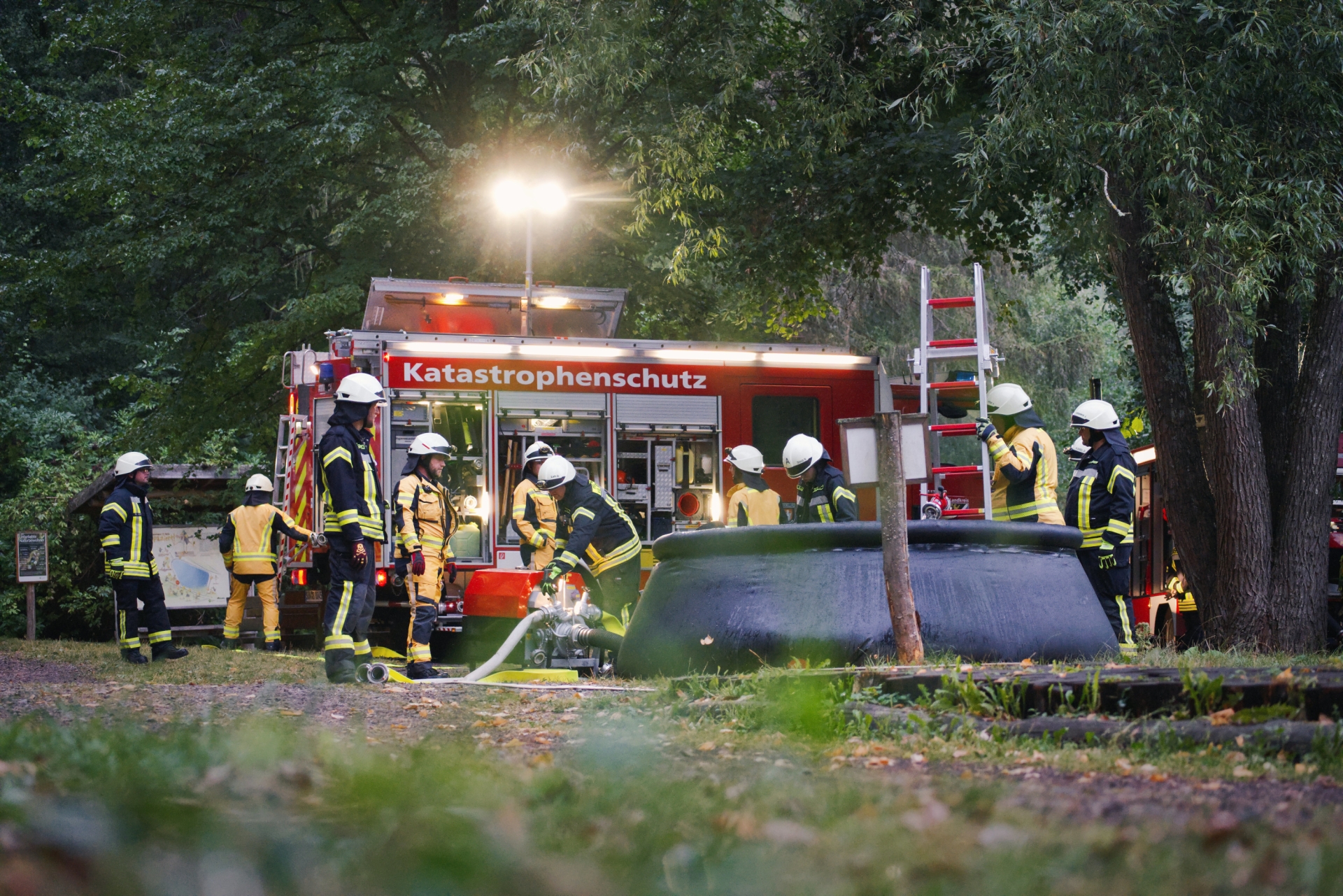 Einsatzkräfte des Katastrophenschutzzugs stehen bei einer Übung an einem roten Einsatzfahrzeug. Im Vordergrund befindet sich ein großes Wasserbacken mit einer Leiter.