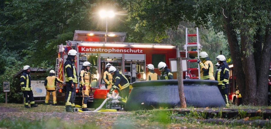 Einsatzkräfte des Katastrophenschutzzugs üben im Wald bei St. Martin die Wasserversorgung bei einem angenommenen Waldbrand. Foto: Tim Domnick Einsatzkräfte des Katastrophenschutzzugs stehen bei einer Übung an einem roten Einsatzfahrzeug. Im Vordergrund befindet sich ein großes Wasserbacken mit einer Leiter.
