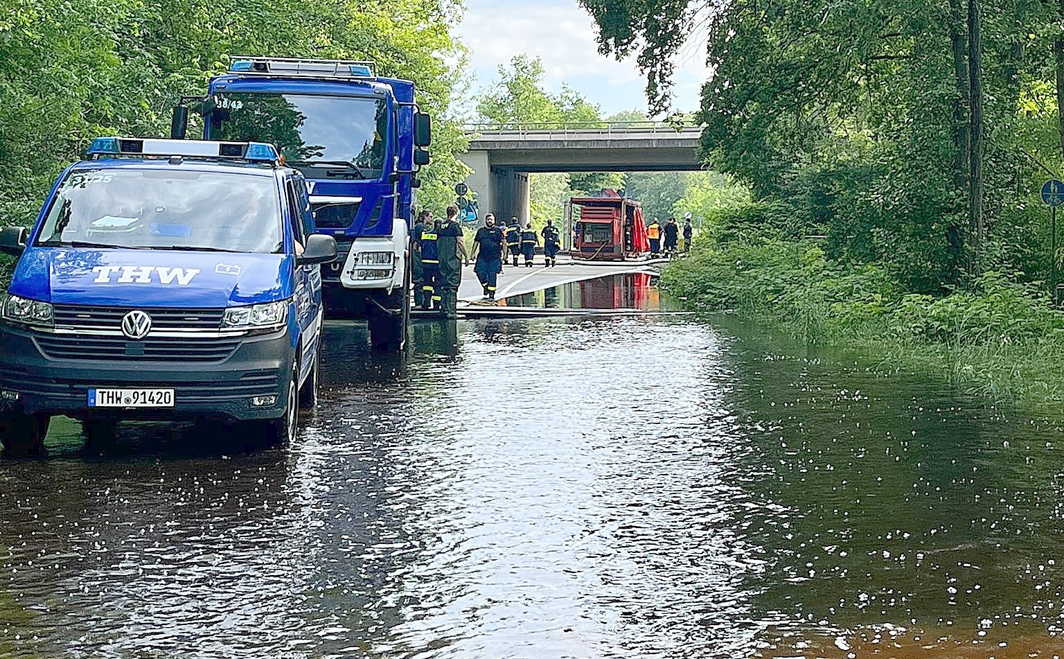 Einsatzkräfte von THW und Feuerwehr stehen mit mehreren Fahrzeugen auf einer überfluteten Straße unter einer Brücke. Das Wasser bedeckt einen großen Teil der Fahrbahn.