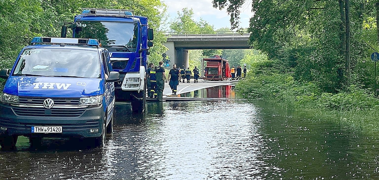 Einsatzkräfte von THW und Feuerwehr stehen mit mehreren Fahrzeugen auf einer überfluteten Straße unter einer Brücke. Das Wasser bedeckt einen großen Teil der Fahrbahn.