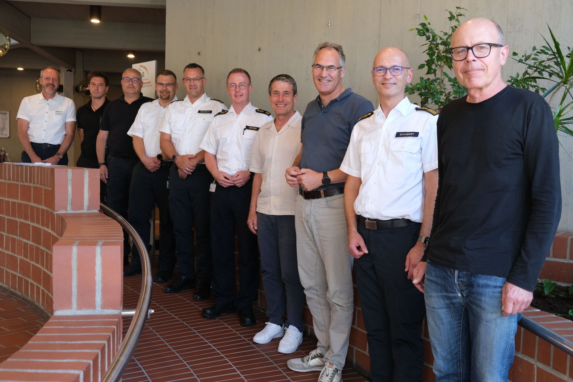 Eine Gruppe von zehn Männern steht für ein Foto in einem Gebäude. Mehrere von ihnen tragen weiße Uniformhemden mit Schulterklappen, die übrigen Freizeitkleidung.