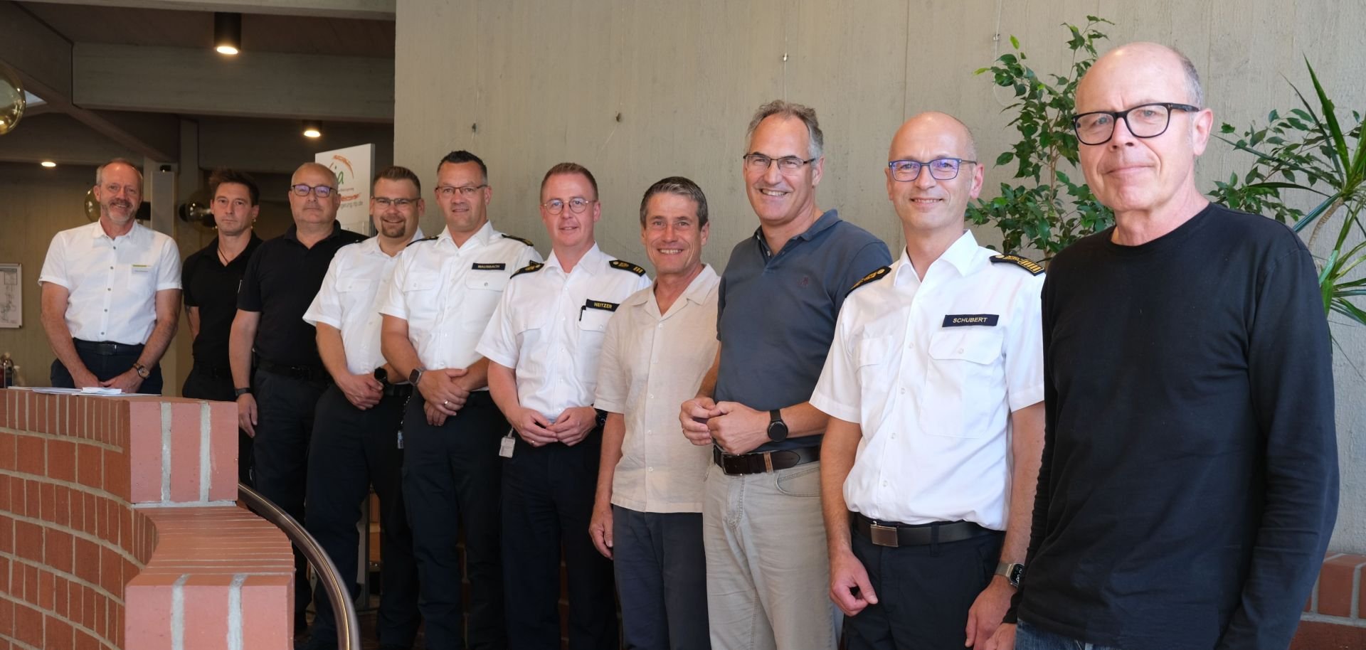 Eine Gruppe von zehn Männern steht für ein Foto in einem Gebäude. Mehrere von ihnen tragen weiße Uniformhemden mit Schulterklappen, die übrigen Freizeitkleidung.