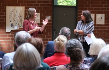 Künstlerin Natascha Brändli (links) erläuterte im Gespräch mit Barbara Auer, worauf es ihr bei ihren künstlerischen Werken ankommt. Foto: KV SÜW Eine Frau spricht vor einem sitzenden Publikum und wendet sich dabei an eine weitere Frau, die neben ihr steht.
