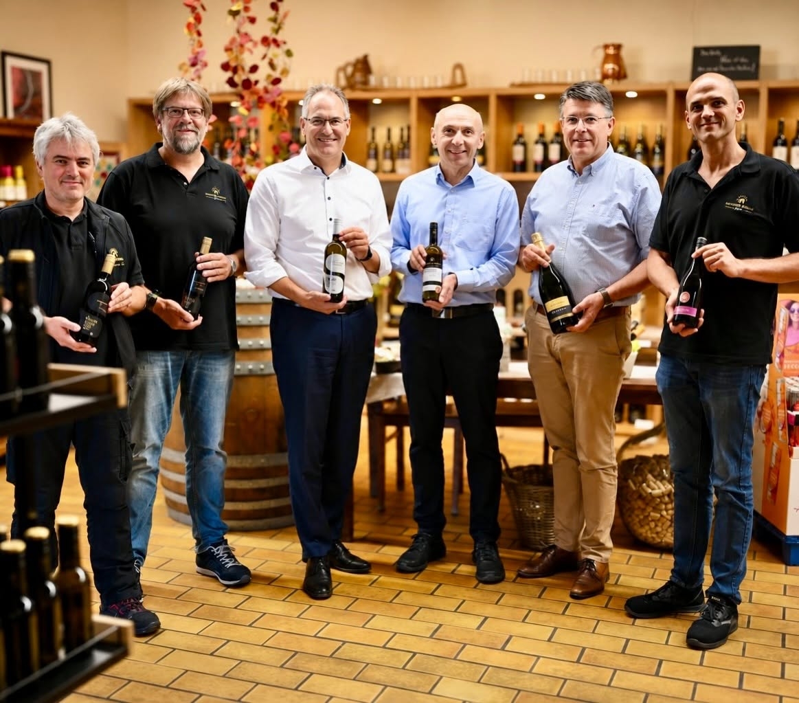 Sechs Männer stehen in einer Vinothek nebeneinander und präsentieren Wein- und Sektflaschen. Im Hintergrund sind Regale mit zahlreichen weiteren Flaschen zu sehen.