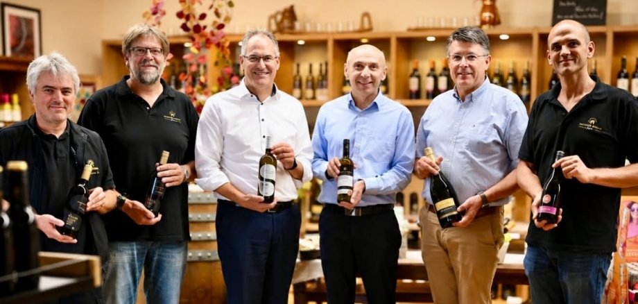 Beim traditionellen Weinlesegespräch bei Deutsches Weintor tauschten sich Vertreter der Genossenschaft mit Landrat Dietmar Seefeldt und Bürgermeister Torsten Blank über die aktuelle Weinlese, die Aussichten für den neuen Jahrgang und die Herausforderungen am Weinmarkt aus. Foto: Deutsches Weintor eG Sechs Männer stehen in einer Vinothek nebeneinander und präsentieren Wein- und Sektflaschen. Im Hintergrund sind Regale mit zahlreichen weiteren Flaschen zu sehen.