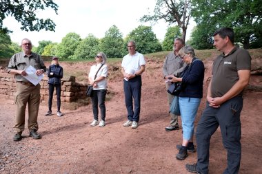 Beim Pressetermin im Wild- und Wanderpark Südliche Weinstraße. Foto: KV SÜW Eine Gruppe steht im Freien an einem bewaldeten Hang. Ein Mann links spricht zu den anderen und hält dabei Unterlagen in den Händen.