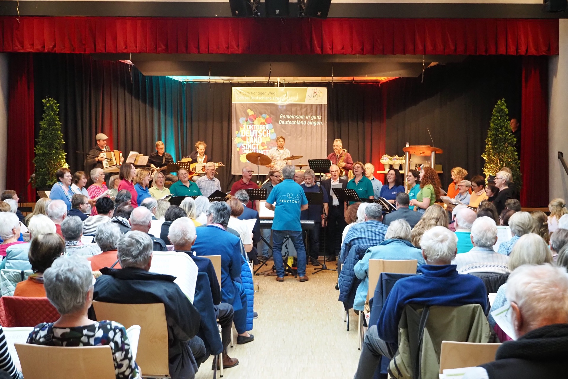 Ein Chor singt auf einer Bühne vor Publikum. Ein Dirigent steht vor der Gruppe, im Hintergrund gebleiten mehrere Musikerinnen und Musiker den Auftritt mit Instrumenten.