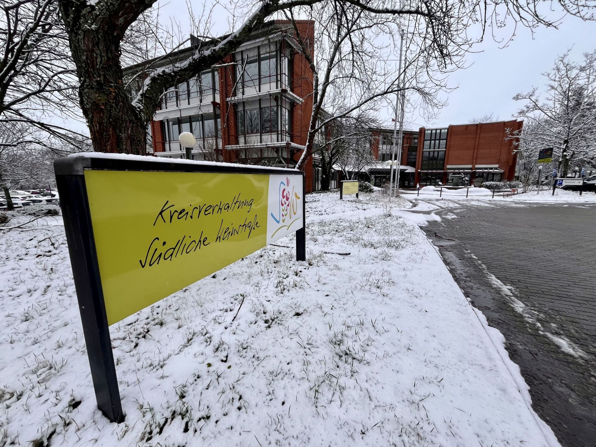 Im Vordergrund ein Schild mit dem Logo und der Aufschrift Kreisverwaltung Südliche Weinstraße, im Hintergrund das verschneite Kreishaus und der Vorplatz mit Brunnen.