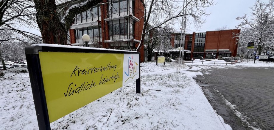 Im Vordergrund ein Schild mit dem Logo und der Aufschrift Kreisverwaltung Südliche Weinstraße, im Hintergrund das verschneite Kreishaus und der Vorplatz mit Brunnen.