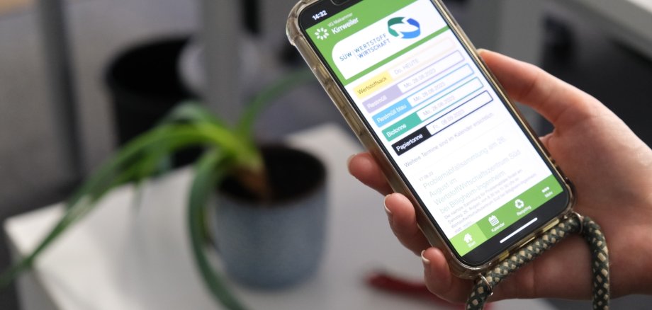 Die SÜW-Wertstoff-App gibt es kostenfrei in den App-Stores. Foto: KV SÜW Eine Hand hält ein Smartphone. Auf dessen Bildschirm wird die SÜW-Wertstoff-App angezeigt. Im Hintergrund ist unscharf eine Zimmerpflanze auf einem Schreibtisch zu sehen.