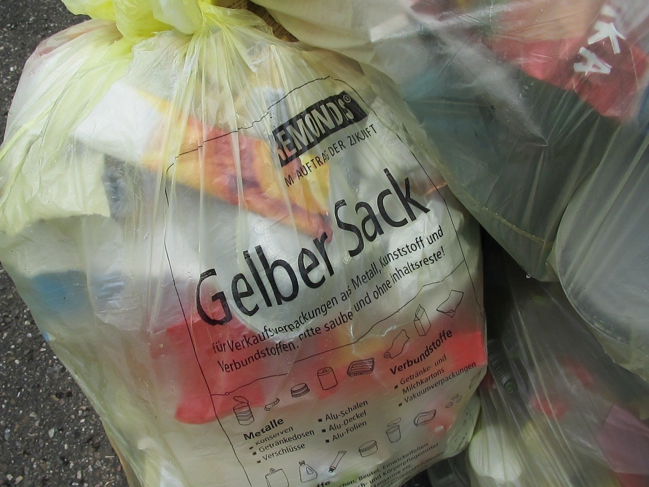 Ein mit Leichtverpackungen gefüllter gelber Sack lehnt an eine Hauswand. 