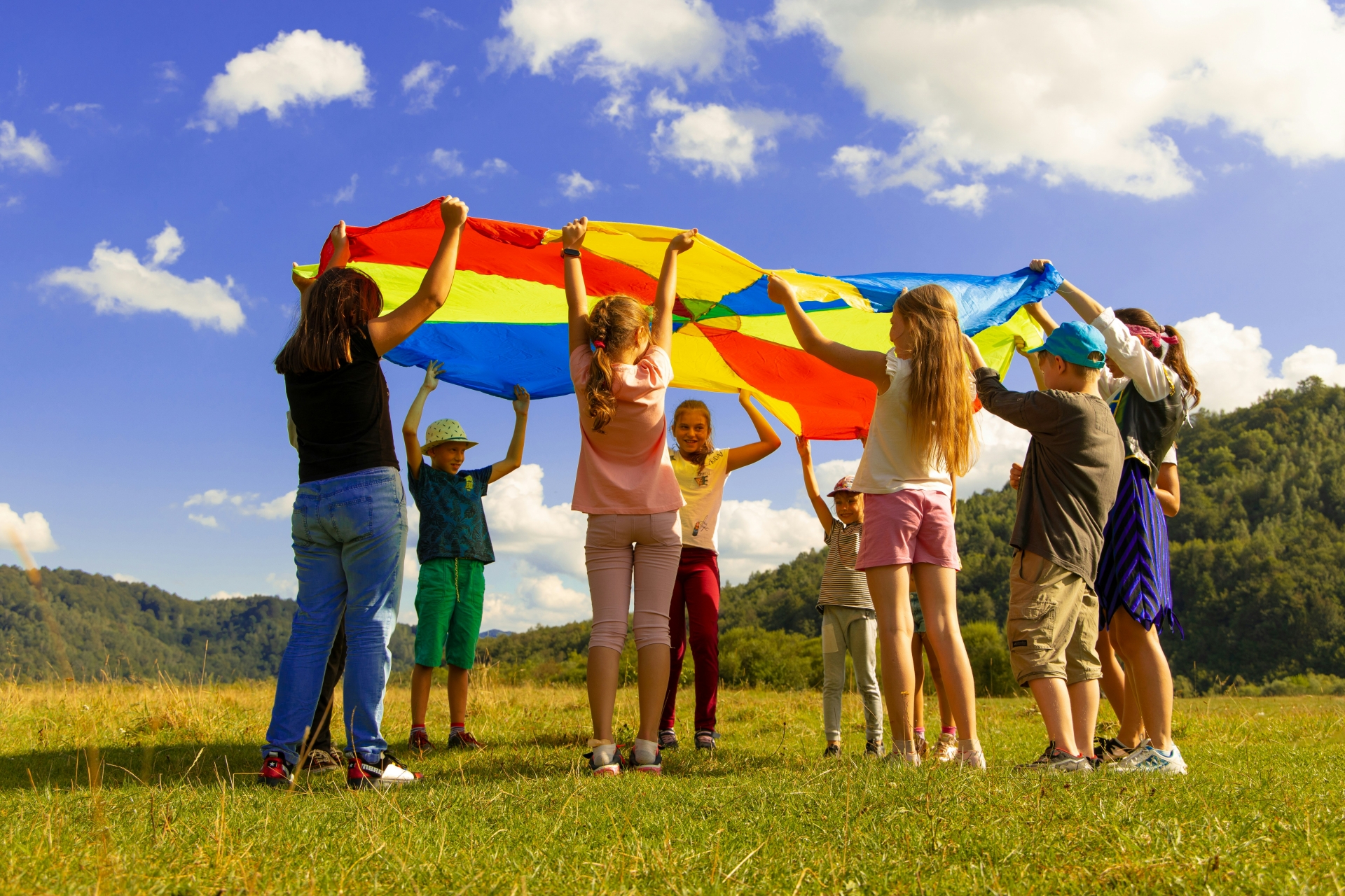 Eine Gruppe von Kindern steht auf einer Wiese im Kreis und hebt gemeinsam ein großes, buntes Schwungtuch in die Luft. Die Szene spielt sich unter einem strahlend blauen Himmel mit einigen weißen Wolken ab. Im Hintergrund sind bewaldete Hügel zu sehen – ein fröhliches Outdoor-Spiel an einem sonnigen Tag in der Natur.