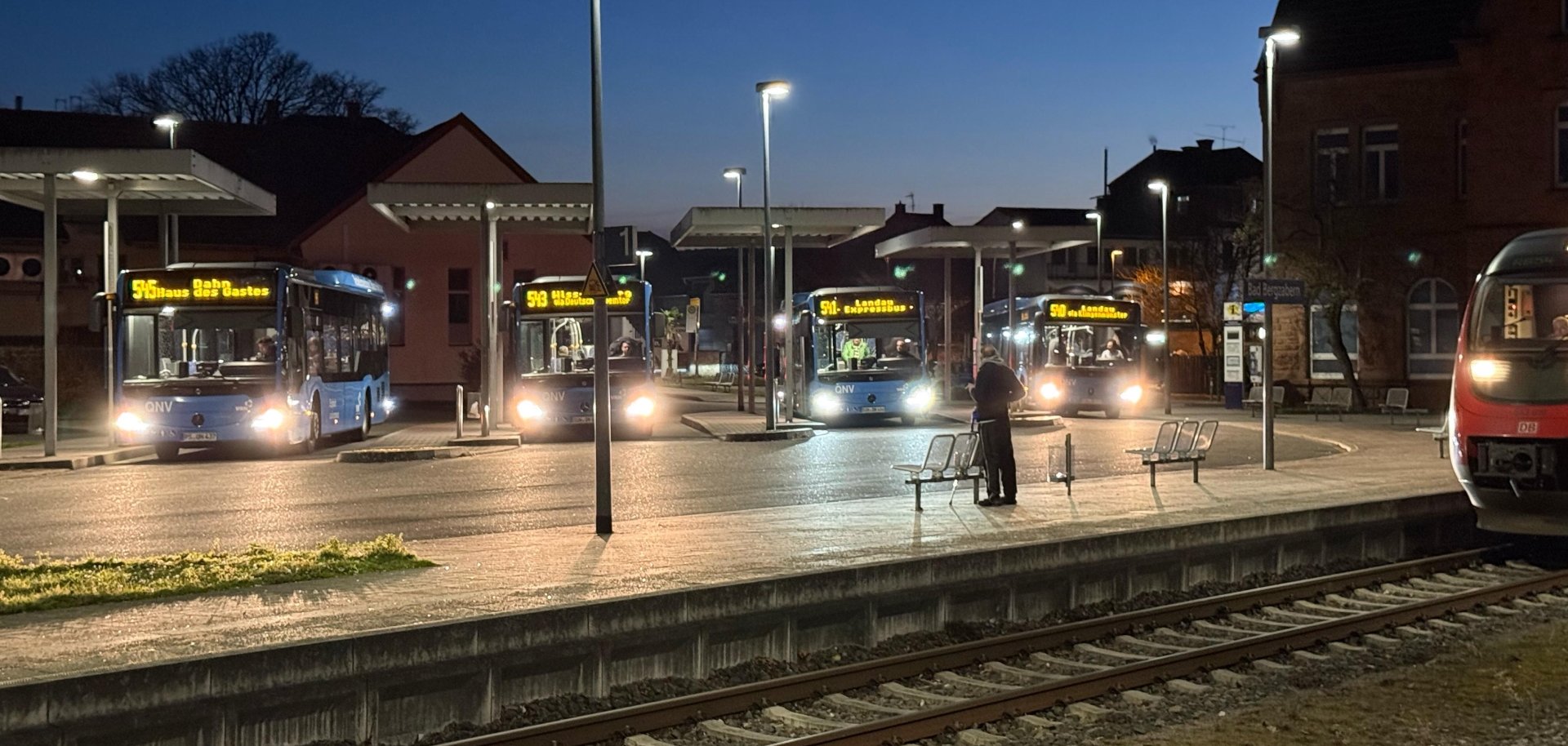 Abendszenerie: Im Vordergrund Schienen und eine Bahn, im Hintergrund vier Busse, die in einem Busbahnhof stehen.