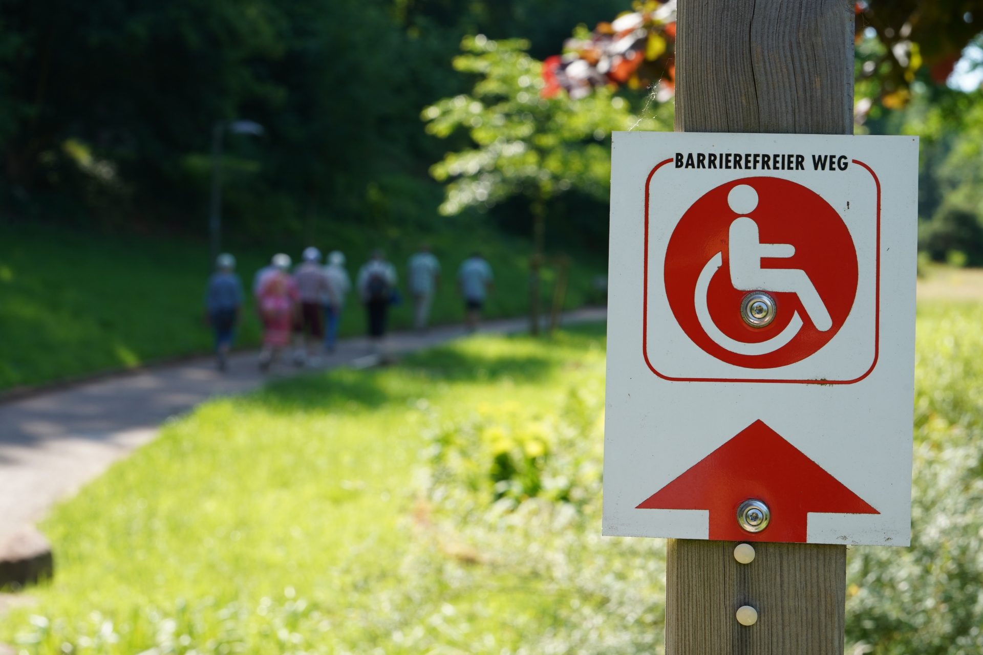 Im Vordergrund ist ein Schild mit einem Piktogramm, auf dem "Barrierefreier Weg" steht, im unscharf gehaltenen Hintergrund laufen auf einem Weg neben einer grünen Rasenfläche mehrere Senioren.