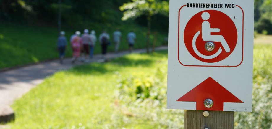Die Markward-Anlage in Annweiler lädt zum Spazieren und Verweilen ein, der Rundweg ist barrierefrei. Foto: KV SÜW Im Vordergrund ist ein Schild mit einem Piktogramm, auf dem "Barrierefreier Weg" steht, im unscharf gehaltenen Hintergrund laufen auf einem Weg neben einer grünen Rasenfläche mehrere Senioren.