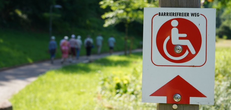 Die Markward-Anlage in Annweiler lädt zum Spazieren und Verweilen ein, der Rundweg ist barrierefrei. Foto: KV SÜW Im Vordergrund ist ein Schild mit einem Piktogramm, auf dem "Barrierefreier Weg" steht, im unscharf gehaltenen Hintergrund laufen auf einem Weg neben einer grünen Rasenfläche mehrere Senioren.