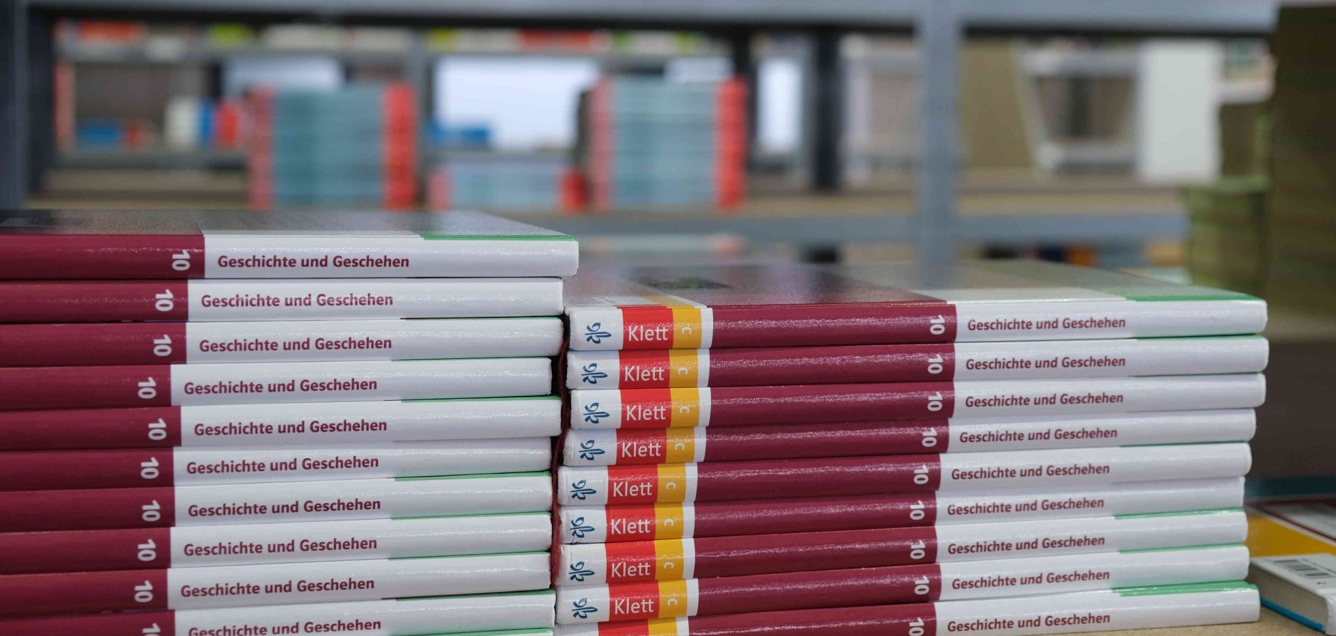 Gestapelte Schulbücher mit dem Titel „Geschichte und Geschehen“ liegen auf einem Tisch in einem Lagerraum. Im Hintergrund sind weitere Bücherregale mit Schulbüchern zu sehen.