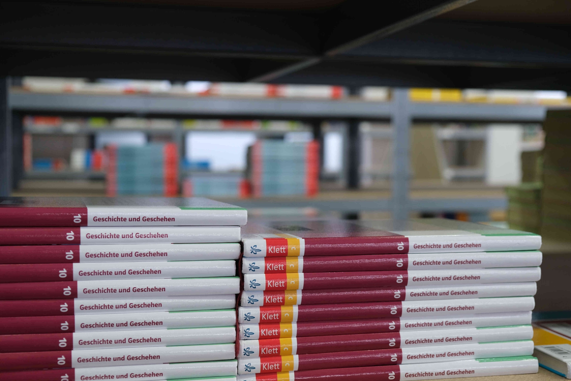 Gestapelte Schulbücher mit dem Titel „Geschichte und Geschehen“ liegen auf einem Tisch in einem Lagerraum. Im Hintergrund sind weitere Bücherregale mit Schulbüchern zu sehen.