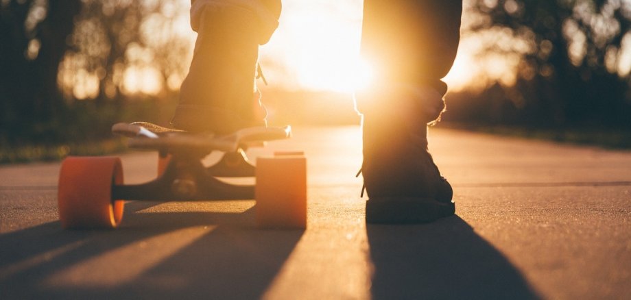 Ein Teenager steht auf einem Skateboard, das von der Abendsonne beleuchtet wird. Die orangefarbenen Rollen und die Silhouette der Person heben sich vor einem warmen, stimmungsvollen Sonnenuntergang ab. (unterstützt von KI)