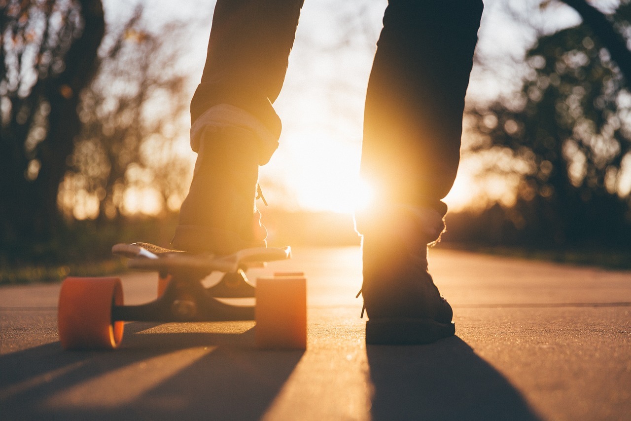 Ein Teenager steht auf einem Skateboard, das von der Abendsonne beleuchtet wird. Die orangefarbenen Rollen und die Silhouette der Person heben sich vor einem warmen, stimmungsvollen Sonnenuntergang ab. (unterstützt von KI)