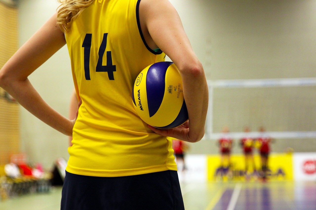 Oberkörper einer Frau im Trikot im Vordergrund, sie trägt einen Volleyball. Im Hintergrund Volleyballnetz und weiter Sportlerinnen.