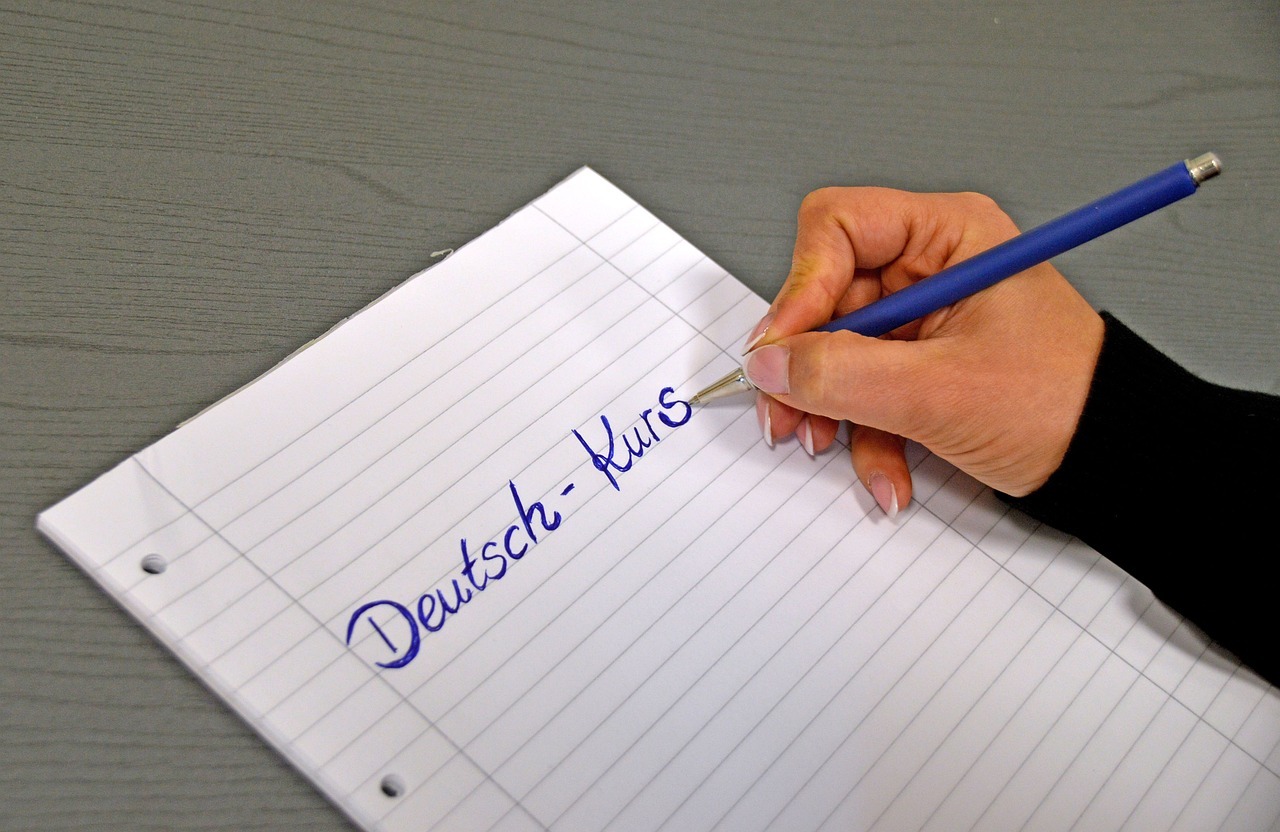 Eine Frau schreibt mit Kugelschreiber den Begriff "Deutsch-Kurs" auf ein Blatt Papier. Foto: Pixabay