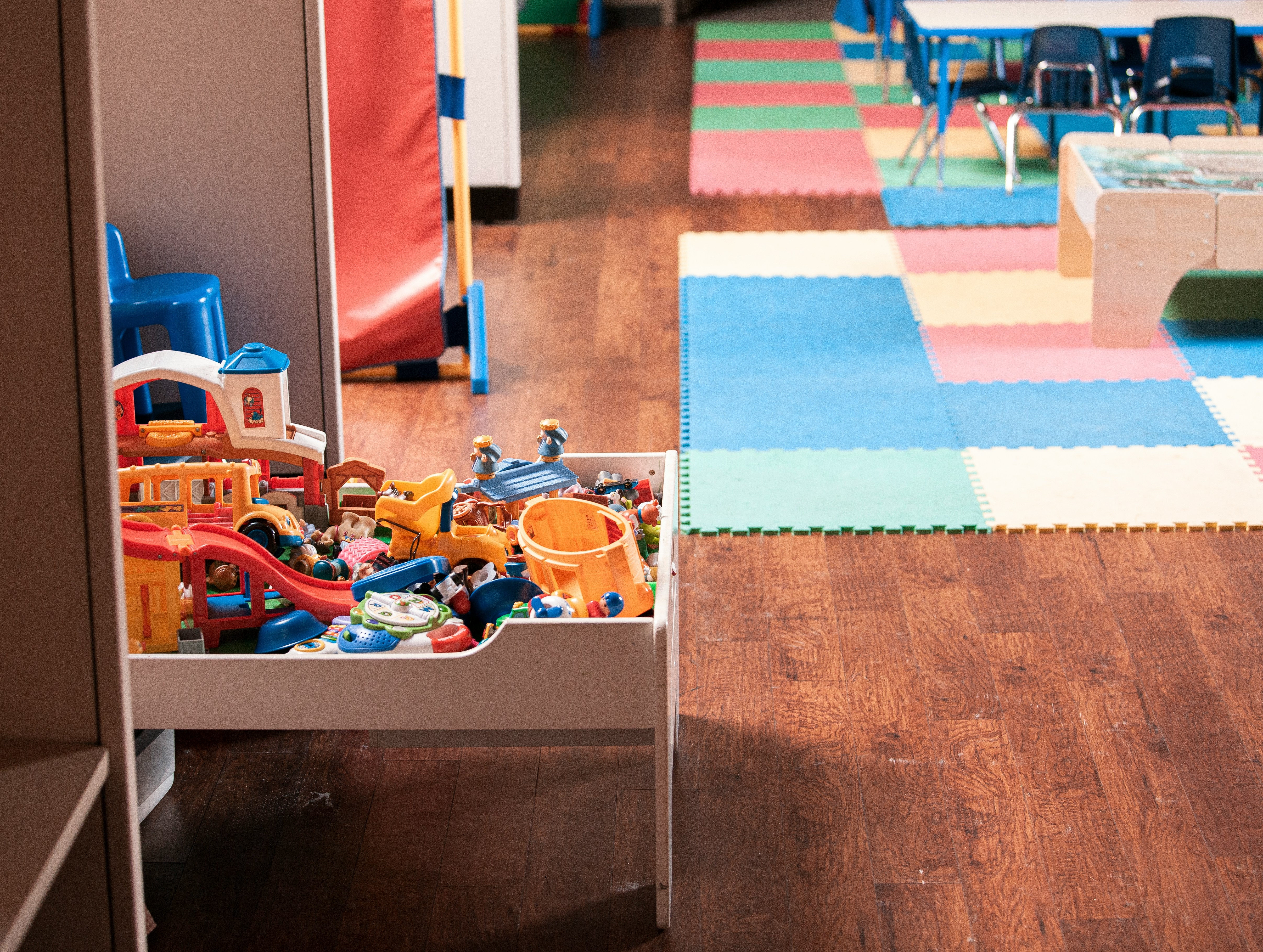 Blick in den Gruppenraum einer Kindertagesstätte. Im Vordergrund steht ein kleiner Tisch mit diversen Kinderspielzuegen darauf, im Hintergrund sind bunte Schaumstoffmatten und eine Sitzgruppe für Kinder zu sehen.