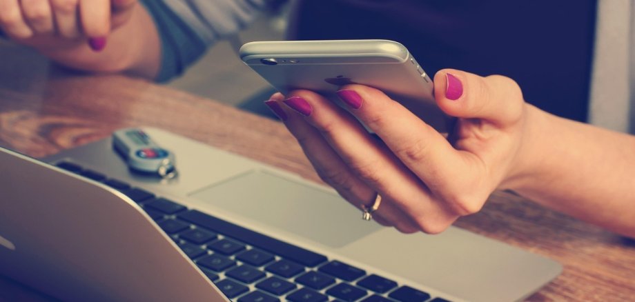 Online-Terminvereinbarung. Symbolfoto: Pixabay Nahaufnahme einer Person mit lackierten Fingernägeln, die ein Smartphone in der Hand hält. Auf dem Tisch liegt ein geöffnetes Notebook mit einem USB-Stick darauf.