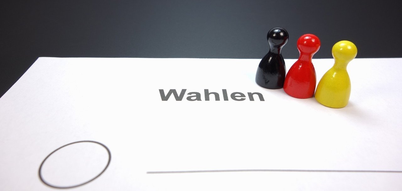 Auf einem Blatt Papier stehen drei Spielfiguren in den Farben schwarz rot und gold. Das Blatt trägt die Überschrift "Wahlen". Darunter ist ein Kreis zum Ankreuzen und eine Linie zu sehen. Es wird somit angedeutet, dass es sich um einen nicht näher definierten Wahlzettel handelt.