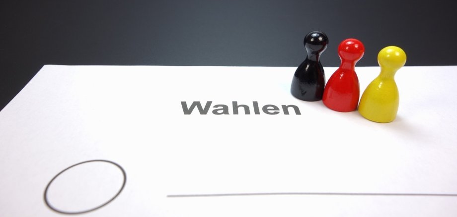 Auf einem Blatt Papier stehen drei Spielfiguren in den Farben schwarz rot und gold. Das Blatt trägt die Überschrift "Wahlen". Darunter ist ein Kreis zum Ankreuzen und eine Linie zu sehen. Es wird somit angedeutet, dass es sich um einen nicht näher definierten Wahlzettel handelt.