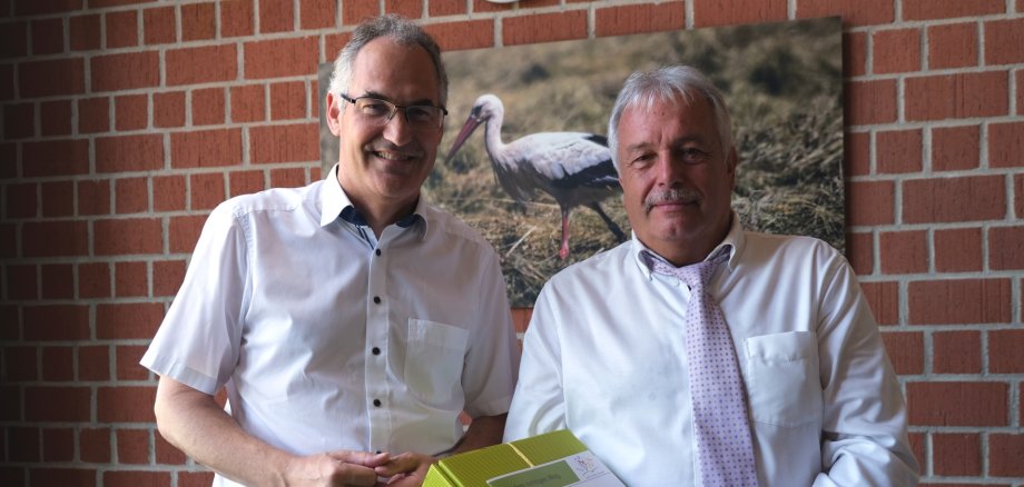 Landrat Dietmar Seefeldt und Rolf Mäckel nebeneinander im Porträt. Rolf Mäckel hält einen grünen Präsentkarton sowie eine Mappe mit Logo und Wappen des Landkreises SÜW in Händen.