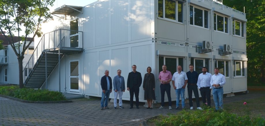 Mehrere Menschen stehen in einer Reihe vor einem Klassenraum-Container auf einem Parkplatz.