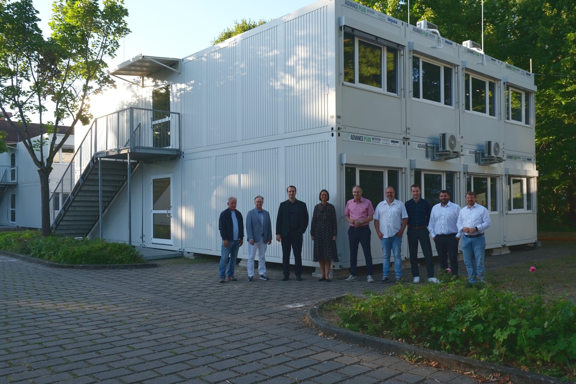 Mehrere Menschen stehen in einer Reihe vor einem Klassenraum-Container auf einem Parkplatz.