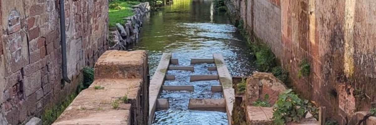Blick auf einen Fluss zwischen zwei Häsuern, im Wasser Betonblöcke, die Stufen bilden.