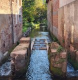 Blick auf einen Fluss zwischen zwei Häsuern, im Wasser Betonblöcke, die Stufen bilden.