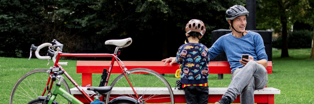 Ein Mann mit Fahrradhelm sitzt auf einer roten Parkbank und schaut auf sein Smartphone. Neben ihm steht ein Kind mit Helm, davor sind ein Kinderfahrrad und ein Rennrad abgestellt, im Hintergrund ist eine grüne Parkanlage zu sehen.
