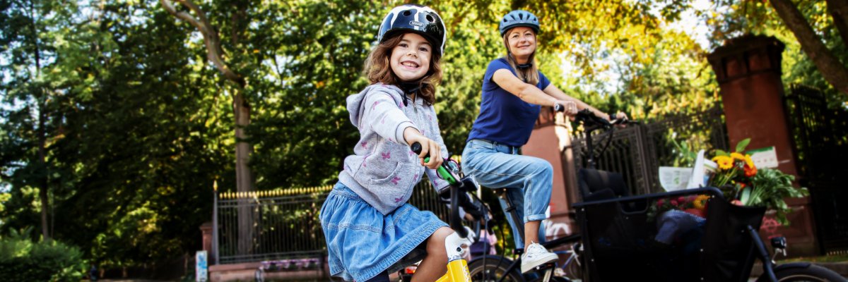 Ein Mädchen fährt mit Helm auf einem gelben Fahrrad über eine Straße und lächelt in die Kamera. Hinter ihr fährt eine Frau mit Helm auf einem Lastenrad durch eine grüne, von Bäumen gesäumte Straße.