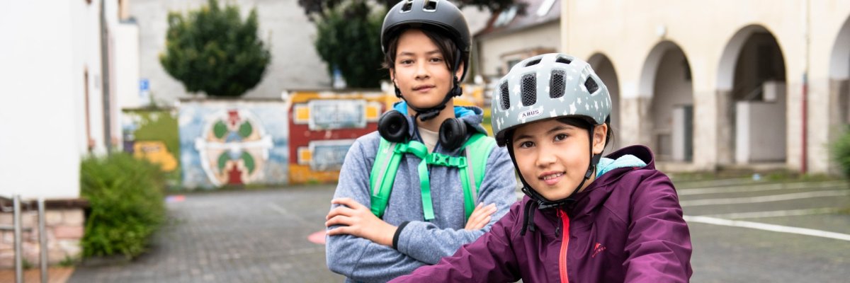 Zwei Kinder mit Fahrradhelmen stehen mit ihren Fahrrädern auf einem gepflasterten Platz vor Gebäuden. Eines sitzt auf dem Fahrrad, das andere steht daneben mit Rucksack und Kopfhörern um den Hals.