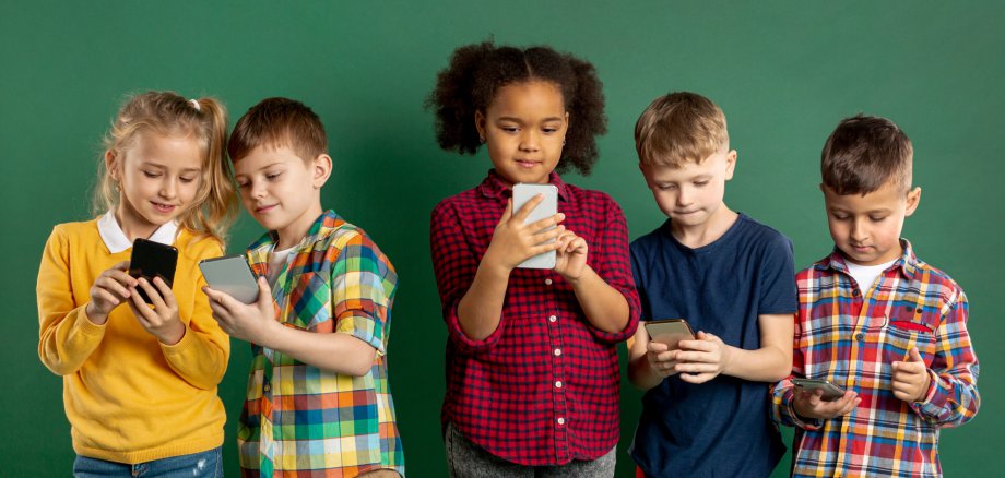 Fünft Kinder stehen nebeneinander vor einem neutralen grünen Hintergrund und schauen jeweils auf ein Smartphone, das sie in Händen halten. Links zeigen sich ein Mädchen und ein Junge gegenseitig etwas auf dem Bildschirm ihres Smarphones.
