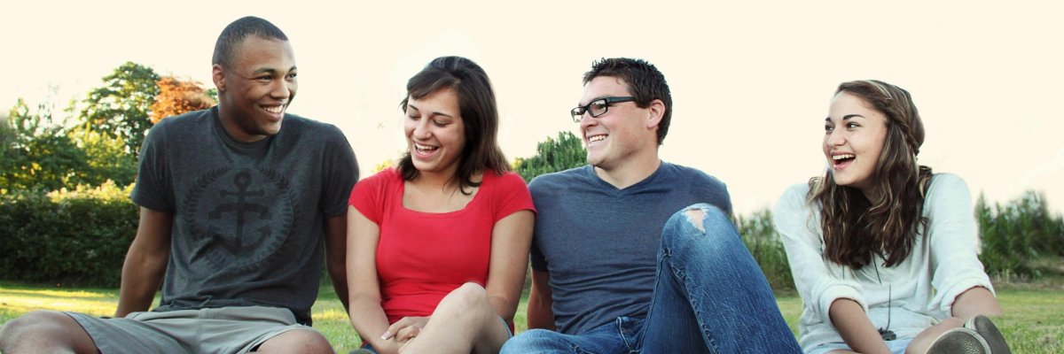 Eine Gruppe von vier jungen Erwachsenen sitzt lachend auf einer Wiese im Freien. Sie wirken entspannt und genießen die gemeinsame Zeit in der Natur.