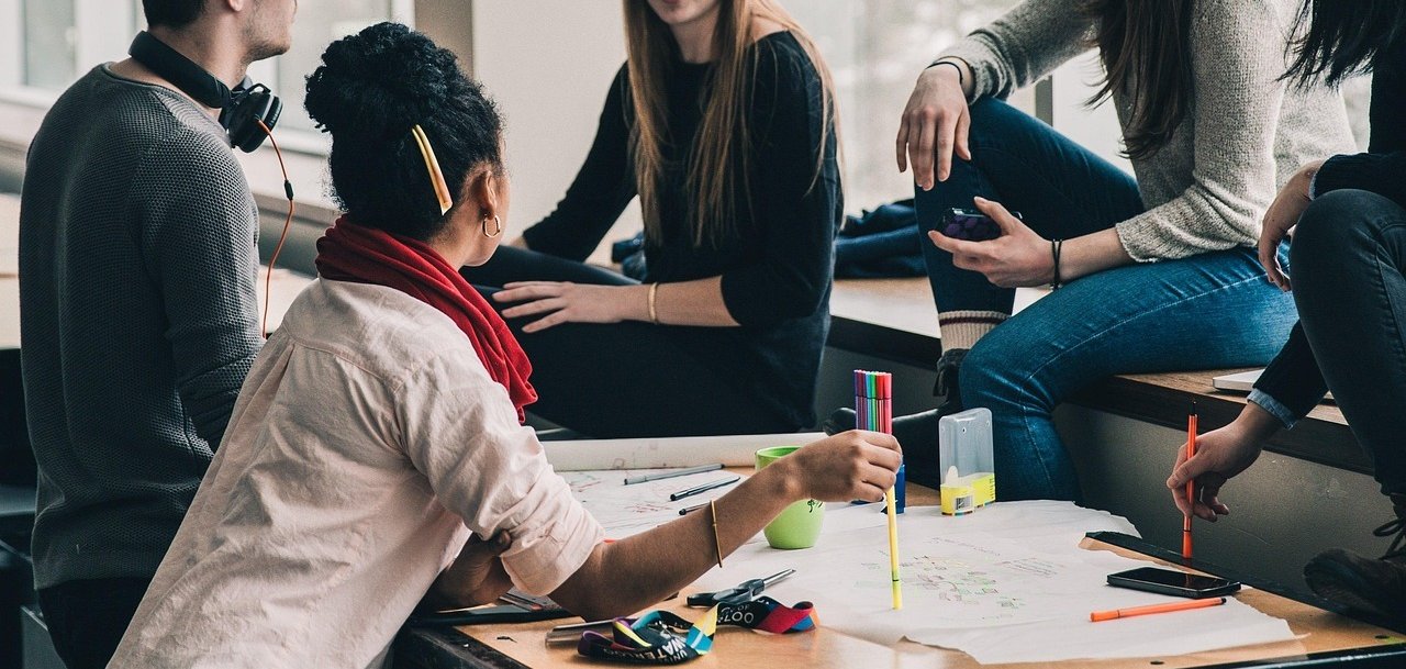 Eine Gruppe junger Menschen sitzt gemeinsam an einem Tisch in einem modernen Raum mit großen Fenstern. Sie arbeiten kreativ an einem Projekt, nutzen bunte Stifte und diskutieren miteinander. Einige sitzen auf der Fensterbank, während eine Person aktiv Notizen auf einem großen Papier macht.