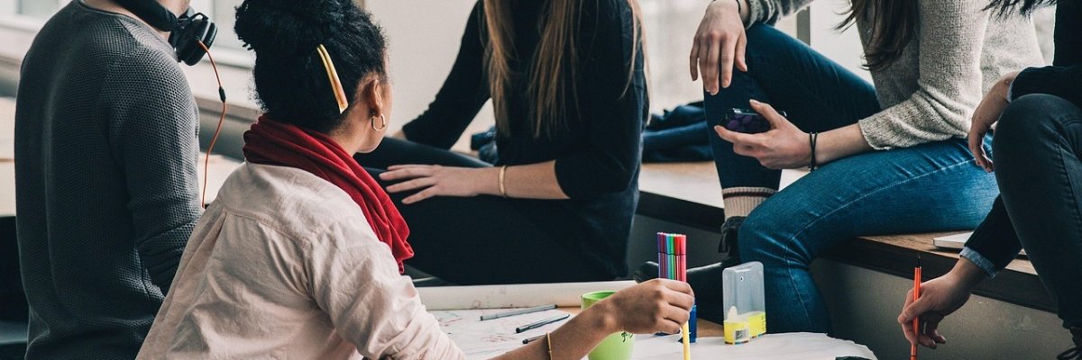 Eine Gruppe junger Menschen sitzt gemeinsam an einem Tisch in einem modernen Raum mit großen Fenstern. Sie arbeiten kreativ an einem Projekt, nutzen bunte Stifte und diskutieren miteinander. Einige sitzen auf der Fensterbank, während eine Person aktiv Notizen auf einem großen Papier macht.