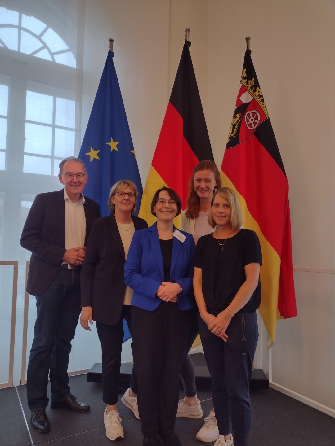 Festakt in der Staatskanzlei Rheinland-Pfalz: Seit zehn Jahren sind die Fachkräfte Gemeindeschwester plus aktiv – der Landkreis SÜW war von Beginn an dabei. Foto: Fünf Personen stehen gemeinsam vor den Flaggen der Europäischen Union, Deutschlands und von Rheinland-Pfalz. Die Gruppe posiert lächelnd für ein Foto bei einer offiziellen Veranstaltung.