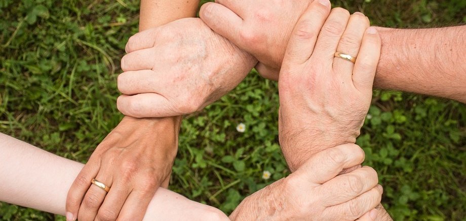 Die Hände mehrerer Personen ergreifen sich gegenseitig am Handgelenk und bilden so einen Kreis als Zeichen der Gemeinschaft.