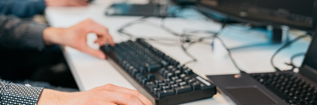 Das Bild zeigt eine Person, die an einem Schreibtisch arbeitet und dabei eine Computermaus und eine Tastatur benutzt. Der Fokus liegt auf den Händen der Person: die rechte Hand bedient die Maus, während die linke Hand auf der Tastatur ruht. Im Hintergrund sind verschwommen mehrere Monitore und weitere Arbeitsplätze zu erkennen.