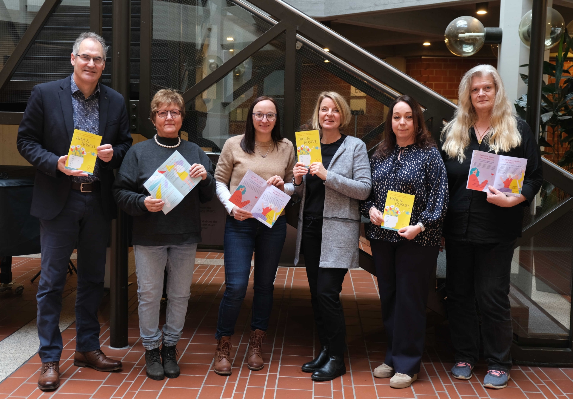Gruppenbild der Gleichstellungsbeauftragten Inge Keller, Nicole Kruppenbacher, Isabelle Stähle, Sandra Hubert und Marita Rothmann mit Landrat Dietmar Seefeldt vor einem Treppenaufgang im Foyer des Kreishauses der Südlichen Weinstraße in Landau. Alle halten ein Programmhaft der Gleichstellungswochen "Brot und Rosen" 2026 in Händen.
